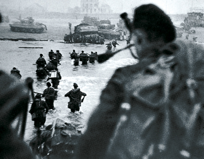 6 juin 1944. Le LCI 519 de Lord Lovat est arrivé devant la plage de Colleville encombrée de chars DD du B Squadron du 13th/18th Hussars face à la maison de la Mer (actuellement entre les rues de Pont l’Évêque et de l’Amiral Courbet). Lord Lovat vient de débarquer (on l’aperçoit à droite de ses hommes) et, au premier plan à droite chargé de son rucksak (sac à dos - terme d’origine allemande), Bill Millin s’apprête à descendre, coiffé de son béret et la cornemuse à la main. (© IWM)