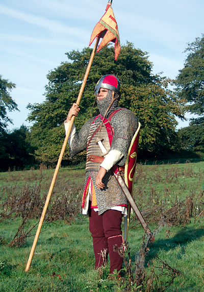 Chevalier normand, capitaine d’un conroi (unité composée de vingt à trente cavaliers) dont il tient la bannière. Il porte des chausses de laine avec jarretières tissées. Il a endossé un haubert de mailles renforcé par un platron lamellaire. Le bouclier est accroché dans le dos. Le casque est à nasal. (© Bertrand Bové)