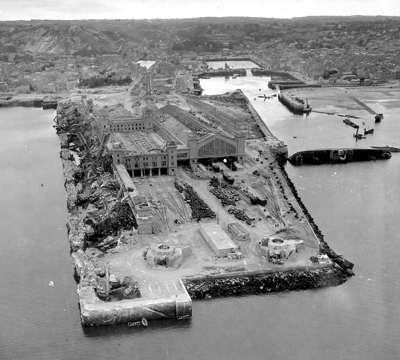 La gare maritime est ravagée par les Allemands. Le quai de France saboté, à gauche, est un amas de ruines. En haut, à gauche, on aperçoit un filet de barrage flottant replié. À droite, le cargo le Normand, sabordé par les Allemands et coulé perpendiculairement à la grande jetée, ne sera renfloué que le 8 novembre 1947. D’ici là, il sert de quai de déchargement. (© National Archives)