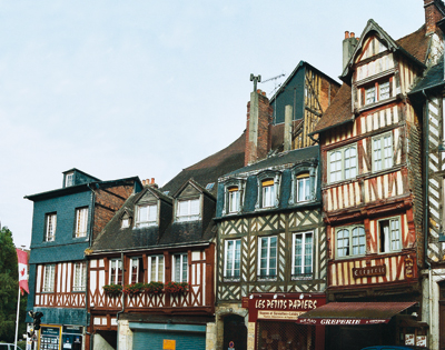 En ayant tourné l’angle de la rue du Maréchal Foch, nous sommes à nouveau dans la Grande Rue où nous retrouvons enfin une série de maisons intactes. De gauche à droite : — A l’angle, le n° 22 présente une maison du xviiie siècle pour ses deux premiers niveaux, qui était en triste état en 1976 (comme l’atteste une photo que nous avions alors faite). Elle a été restaurée et en partie recouverte d’essentes d’ardoises. — Au n° 20 se dresse une jolie maison du xve siècle avec son encorbellement typique. — Suit, au n° 18, une belle maison du xviiie siècle avec l’agencement complexe du pan de bois typique de cette époque. L’étage sous comble a été remanié à la fin du xixe siècle. — Vient ensuite l’une des plus belles maisons de Lisieux, parmi celles qui ont échappé aux destructions. Ce n°16 est une maison en encorbellement du début du xvie siècle. Les deux encorbellements successifs présentent des entretoises avec de remarquables décors de rageurs, typiques de cette époque mais les seuls qui ont survécu aux destructions. Elle aurait pu être amputée d’une troisième travée à droite, au xviiie siècle, quand a été édifiée la maison qu’on distingue sur la droite. Cette belle maison est surmontée d’une lucarne. (EG/PN).