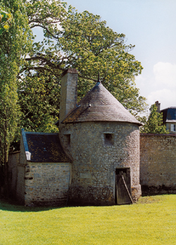 La tour de défense médiévale du château de Damigny vue depuis le nord. Elle est flanquée, à gauche, de son four à pain, et, à droite, d’un vestige du mur de l’enceinte avancée. Au premier étage, elle est encore munie de trois embrasures de tir pour arquebuses. Au rez-de-chaussée, deux d’en­tre elles ont été bouchées, la troisième a disparu lorsqu'une porte a été malencontreusement percée au XIXe siècle. La base de cette tour est légèrement talutée. On remarque la souche de cheminée pour le four à pain et le chauffage de la salle basse, ancienne salle des gardes. (Photo Georges Bernage © Patrimoine Normand)